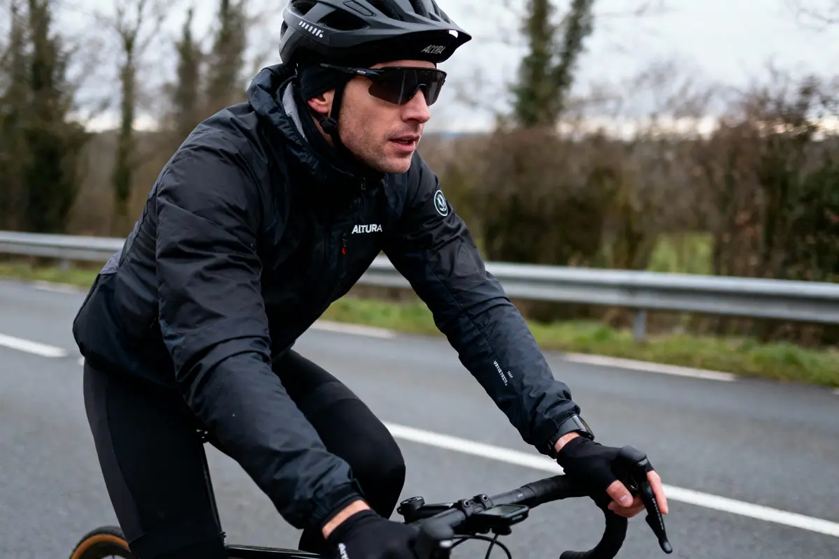 Cycliste en tenue noire, casque et lunettes, pédalant sur route pavée par temps nuageux.