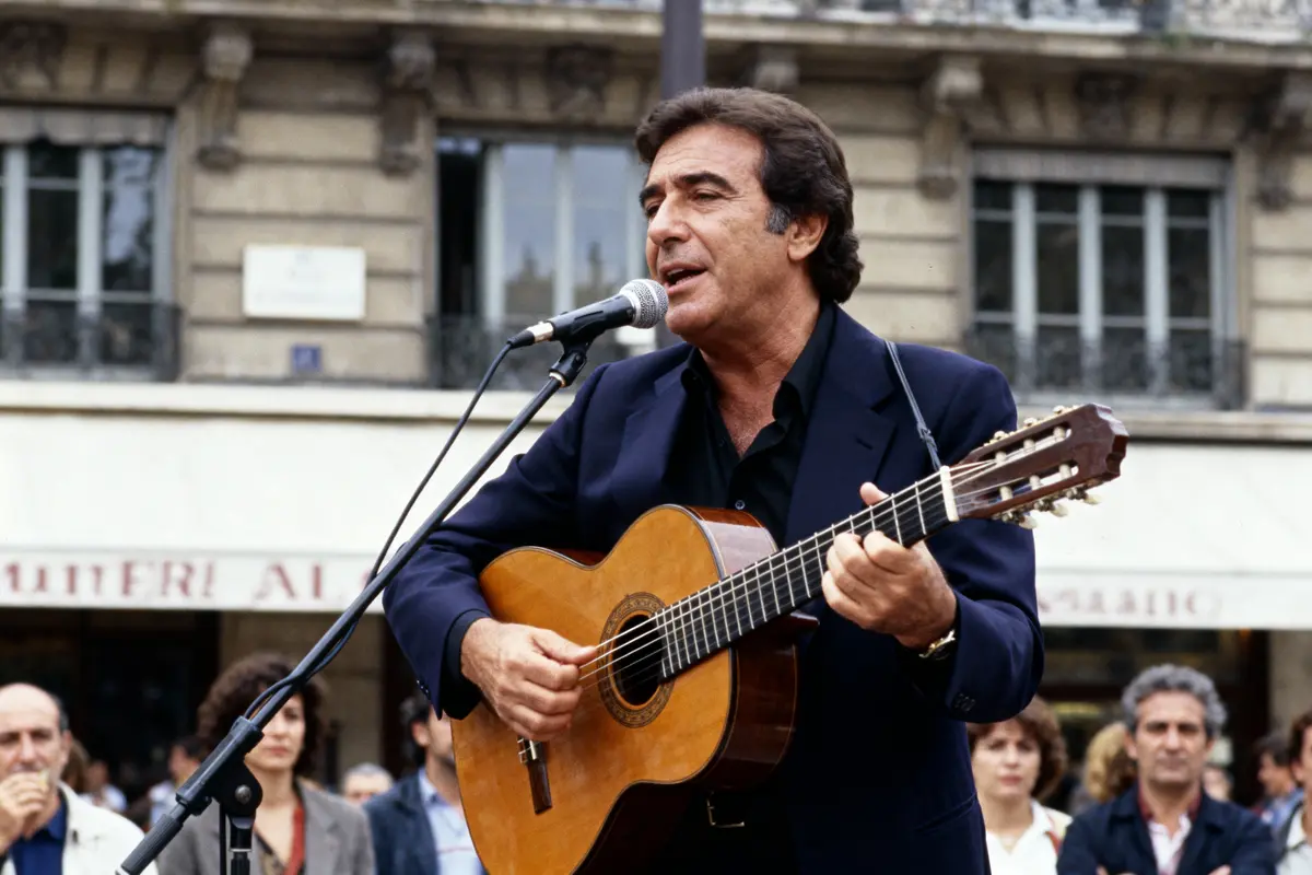 Homme jouant de la guitare acoustique et chantant avec un micro, extérieur devant un immeuble, public en arrière-plan.