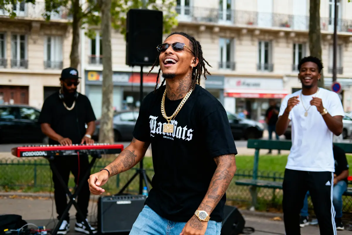 Homme souriant portant lunettes et chaînes en or, devant musicien jouant du clavier en extérieur urbain.