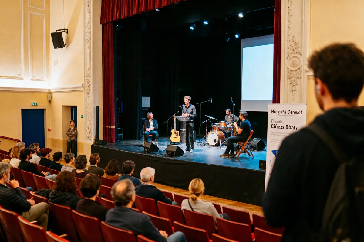 Conférence sur scène avec intervenants, guitariste, batteur devant un public assis dans une salle de spectacle.