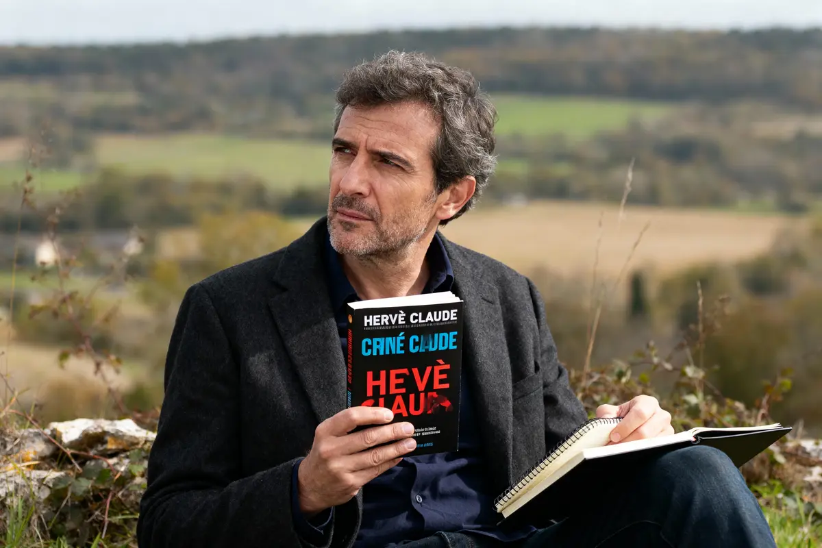 Homme assis tient un livre et un carnet, paysage naturel en arrière-plan, expression pensive, journée ensoleillée.