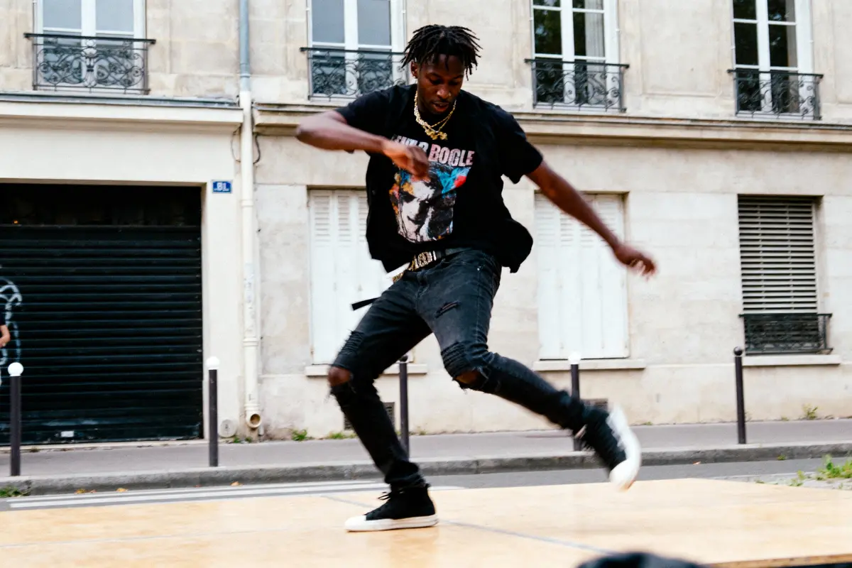 Un homme danse avec énergie dans la rue, portant un t-shirt noir et un jean déchiré.