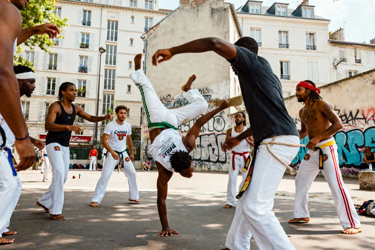 Groupe de personnes pratiquant la capoeira dans une rue, entourées de bâtiments et de graffitis.