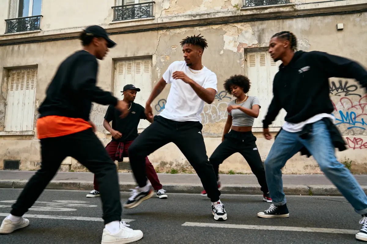Groupe de jeunes dansant énergiquement sur une rue, devant un mur graffité, expression de culture urbaine.