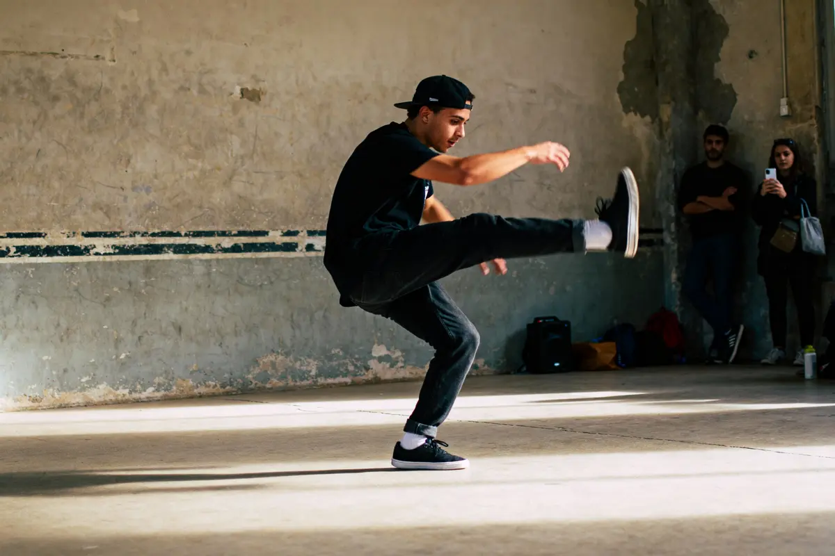 Danseur urbain en mouvement, pied levé, casquette noire, mur béton, spectateurs en arrière-plan.