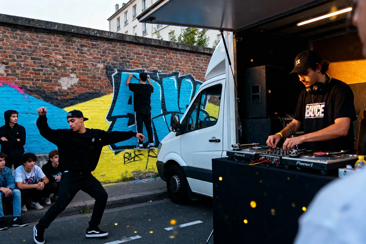 Un DJ mixe, un danseur bouge, et un graffeur peint un mur coloré.
