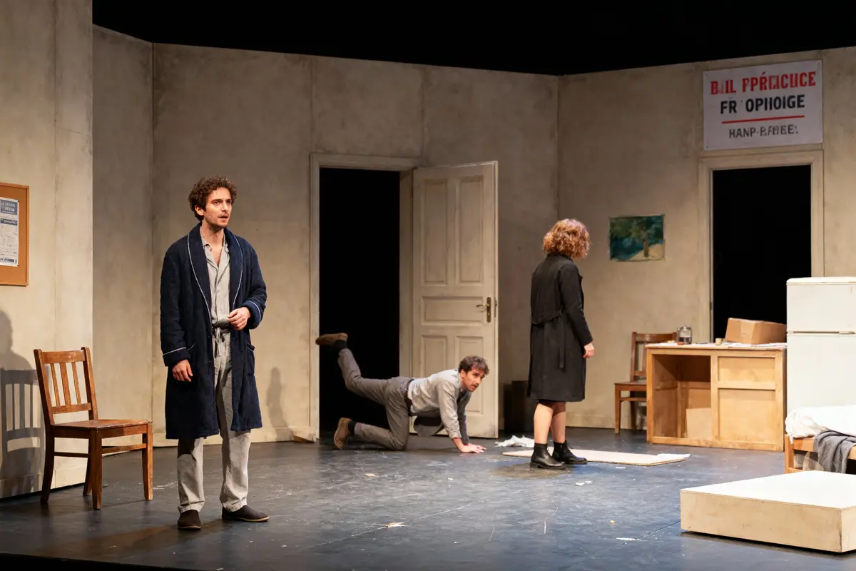 Trois acteurs sur scène dans une pièce, un homme en peignoir, un homme rampant, femme debout.