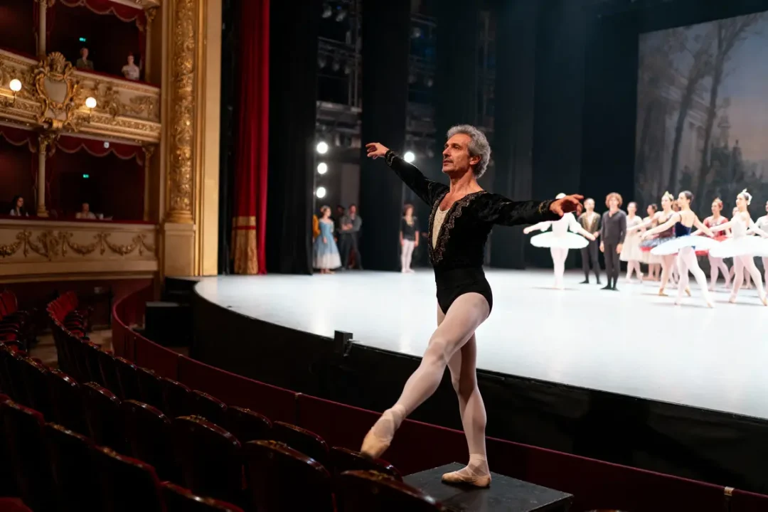 Danseur de ballet en costume noir sur scène, entouré d'un groupe de ballerines, théâtre élégant en arrière-plan.