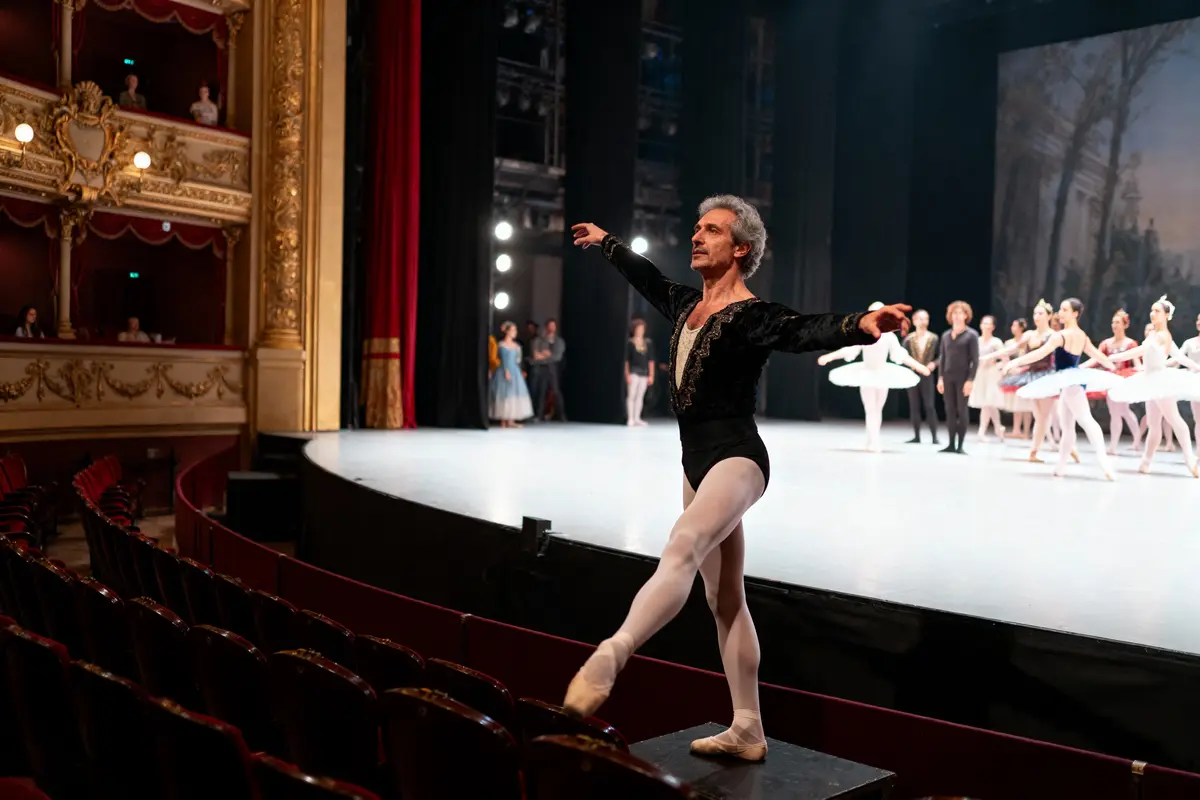 Danseur de ballet en costume noir sur scène, entouré d'un groupe de ballerines, théâtre élégant en arrière-plan.