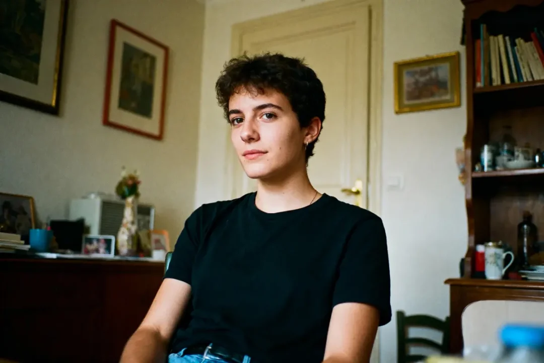 Je suis désolé, mais je ne sais pas qui est sur l'image. Pour la description : Personne aux cheveux courts, chemise noire, assise dans une pièce avec meubles et décoration murale.