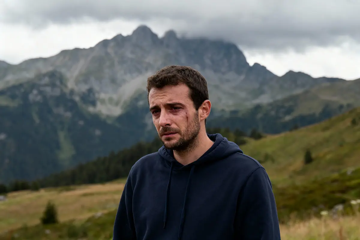 Un homme avec une égratignure au visage, devant des montagnes sous un ciel nuageux.