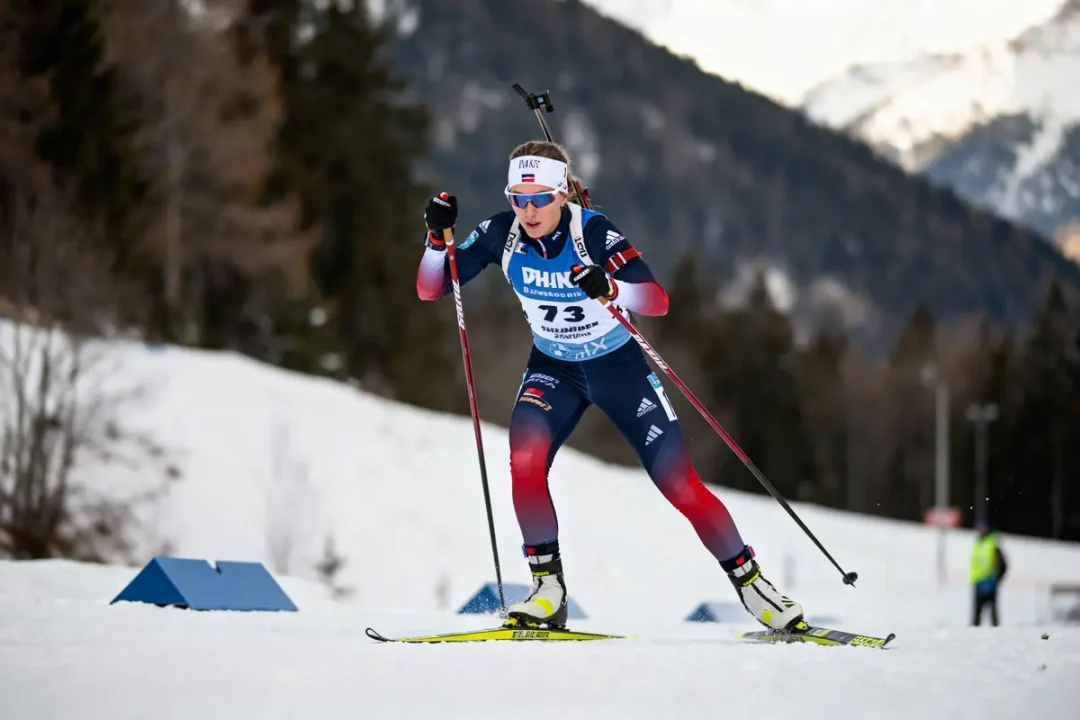 Biathlète en compétition, ski de fond, bâtons, montagne enneigée en arrière-plan, combinaison bleu et rouge.