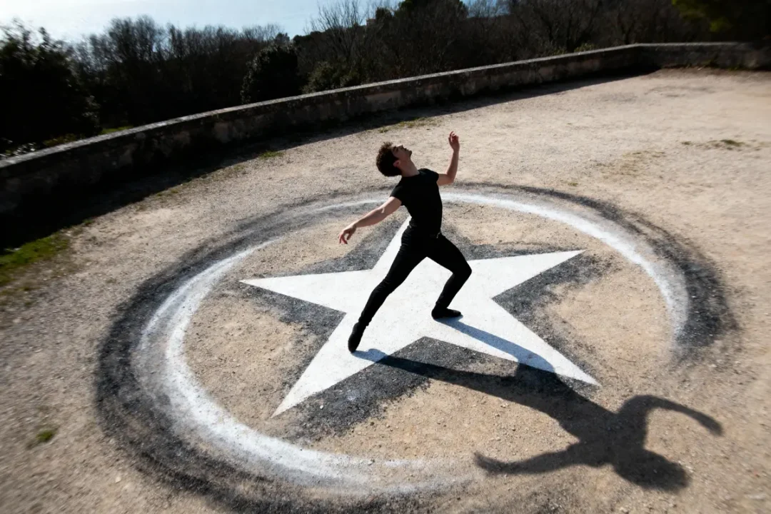 Danseur en noir sur étoile blanche, sol extérieur, ombre dramatique, posture artistique, ciel bleu, arbres en arrière-plan.