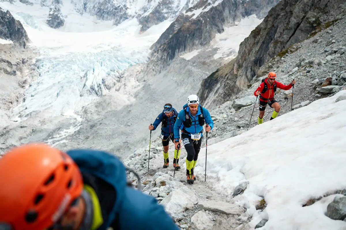 Trois alpinistes escaladent une montagne enneigée avec des bâtons sous un ciel nuageux.