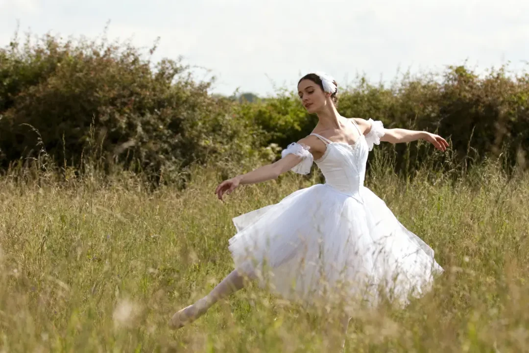 Danseuse en robe blanche dans champ verdoyant, exécutant un mouvement gracieux sous un ciel clair.