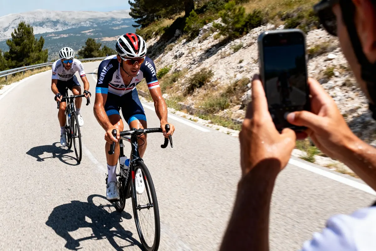 Deux cyclistes montent une route vallonnée, suivis par une personne les photographiant avec un smartphone.