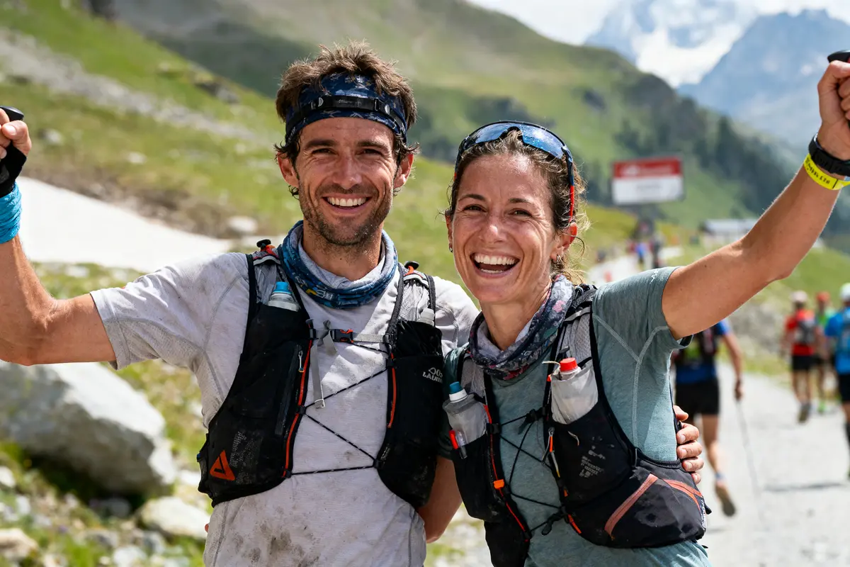Deux personnes souriantes portant des équipements de randonnée, en montagne, avec un paysage alpin en arrière-plan.