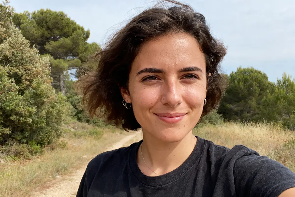 Jeune femme souriante sur un chemin rural, entourée d'arbres et de végétation sous un ciel clair.