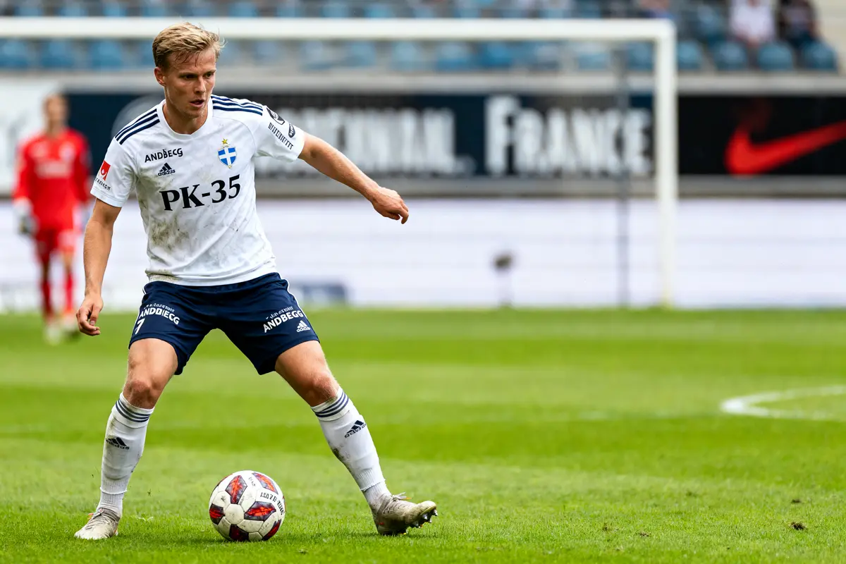 Un joueur de football contrôle le ballon sur un terrain, portant un maillot blanc et bleu PK-35.