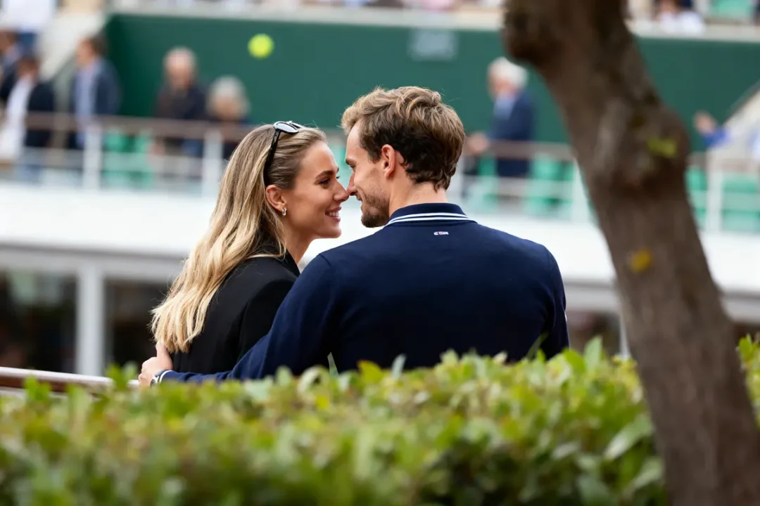 Un couple souriant s'embrasse dans un cadre extérieur, entouré de verdure et de spectateurs flous.
