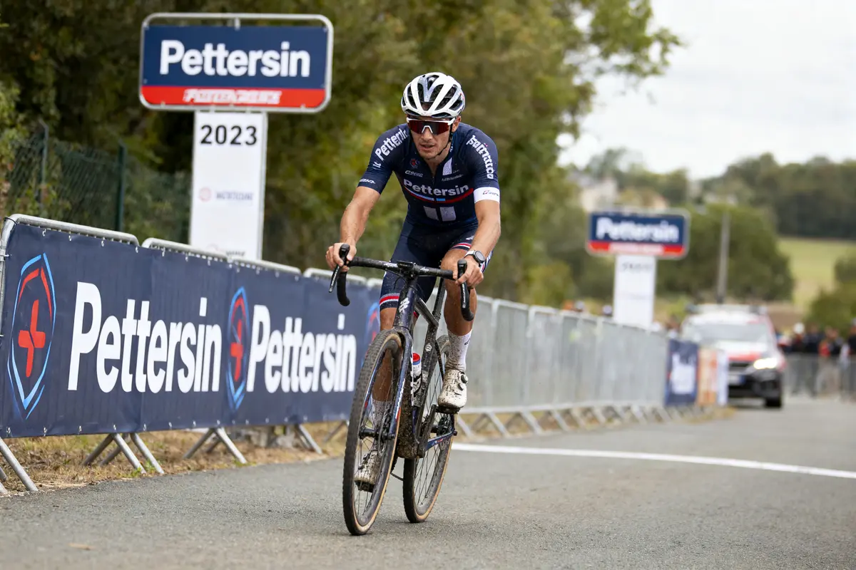 Cycliste en compétition sur route, vêtu de bleu. Bandes "Pettersin" visibles sur panneau et barrière.
