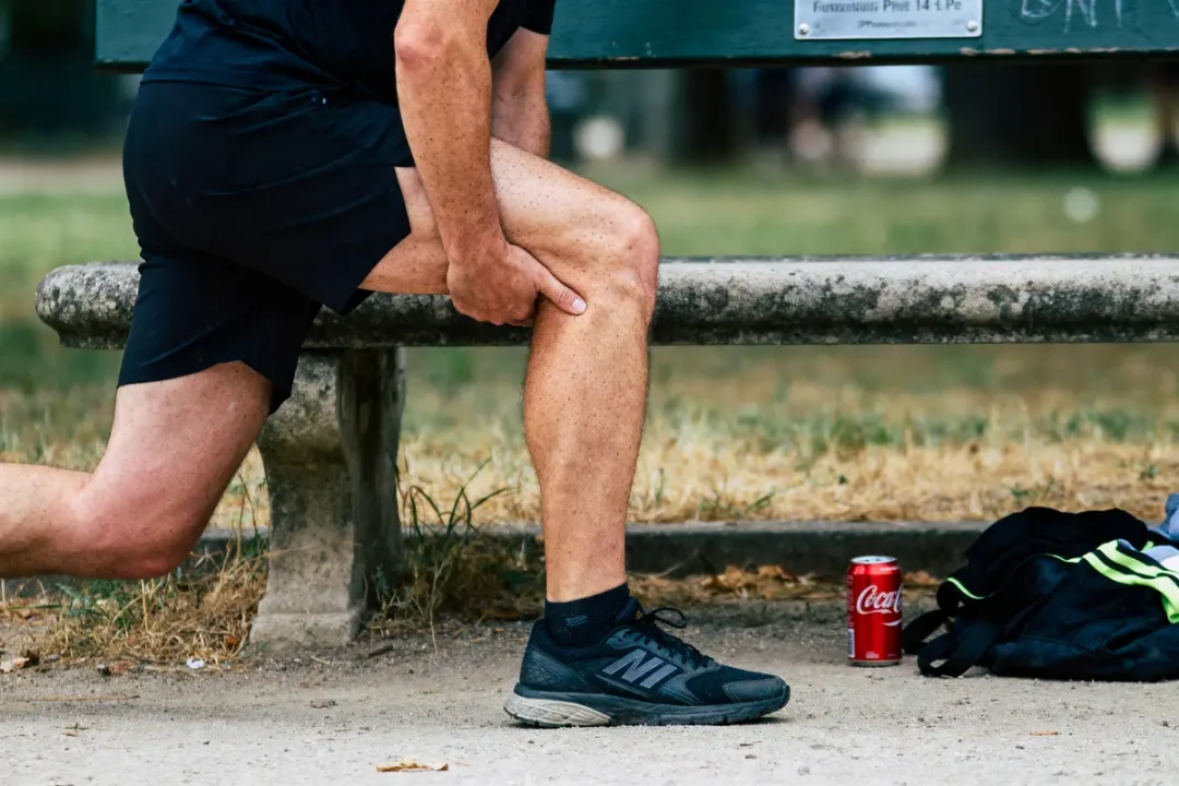 Personne s'étire, main sur mollet, près d'un banc avec canette de soda et sac noir sur sol.