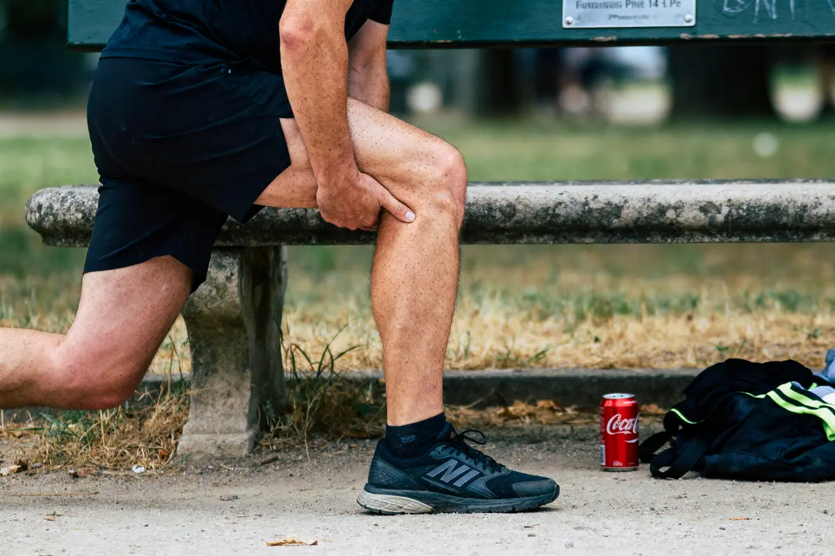 Personne s'étire, main sur mollet, près d'un banc avec canette de soda et sac noir sur sol.