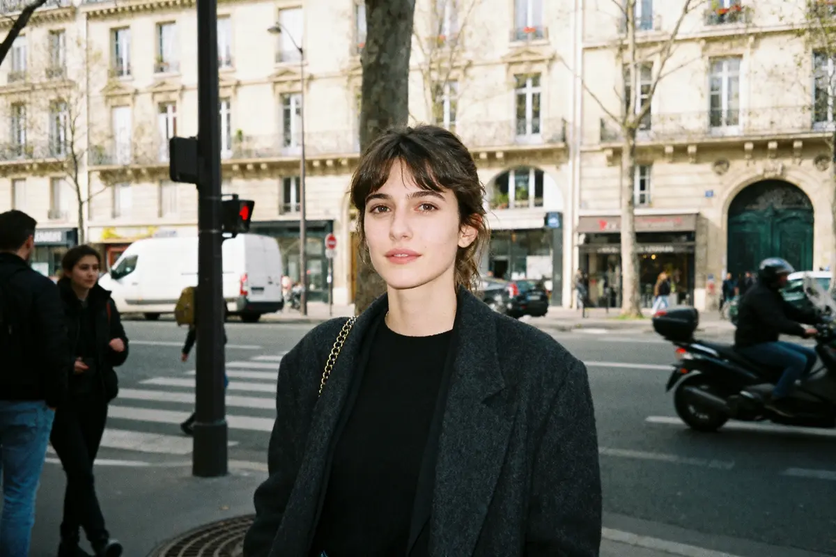 Une femme avec un manteau sombre dans une rue urbaine, bâtiments en arrière-plan, passants et voiture.