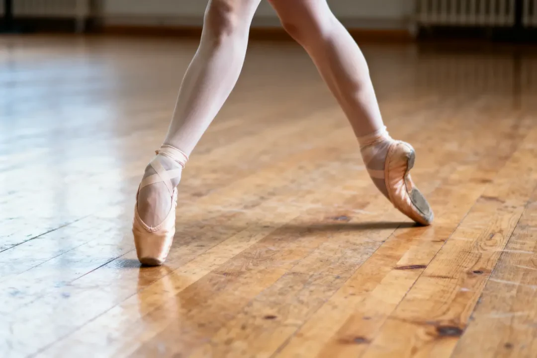 Danseuse en pointe sur parquet, portant des chaussons roses et des collants, salle de danse lumineuse.