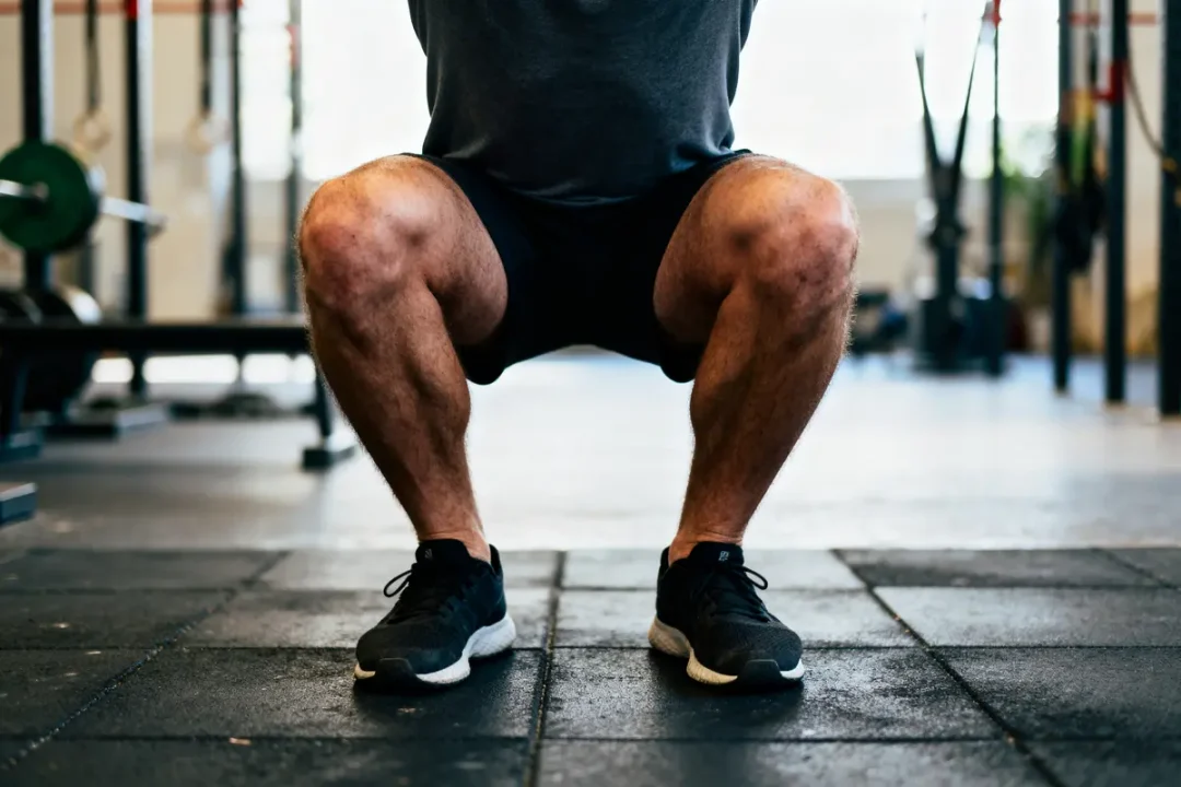 Personne en position accroupie dans une salle de sport, portant des vêtements de sport noirs. Entrainement fitness.