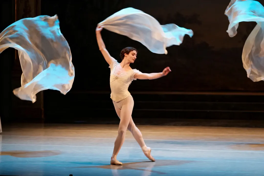 Danseuse de ballet sur scène, costume clair, tenant des voiles flottants, mouvements gracieux, éclairage théâtral doux.