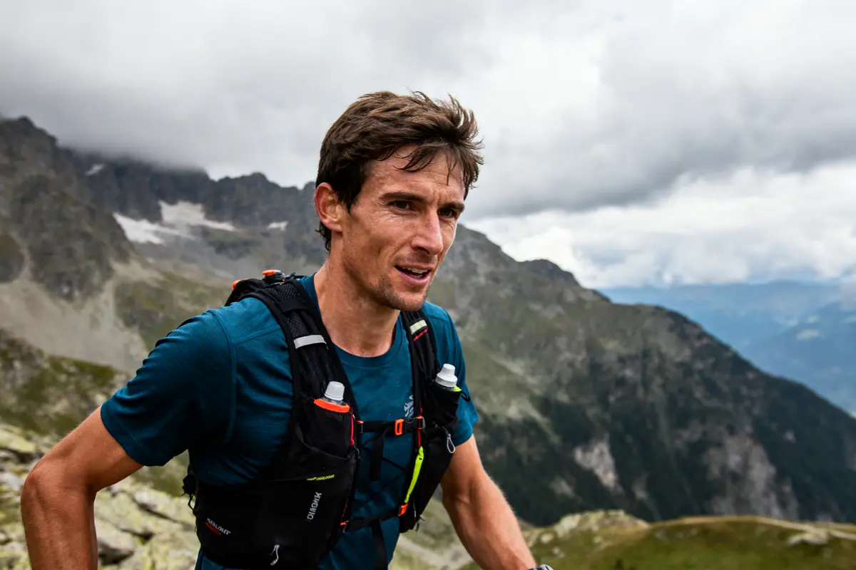 Coureur en montagne avec sac à dos, ciel nuageux, montagnes en arrière-plan.