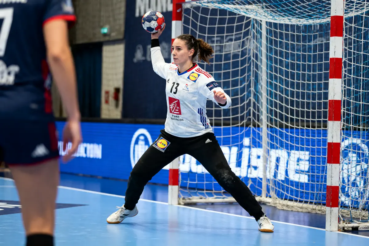 Gardienne de handball en action, prête à stopper un tir, devant un but lors d'un match.