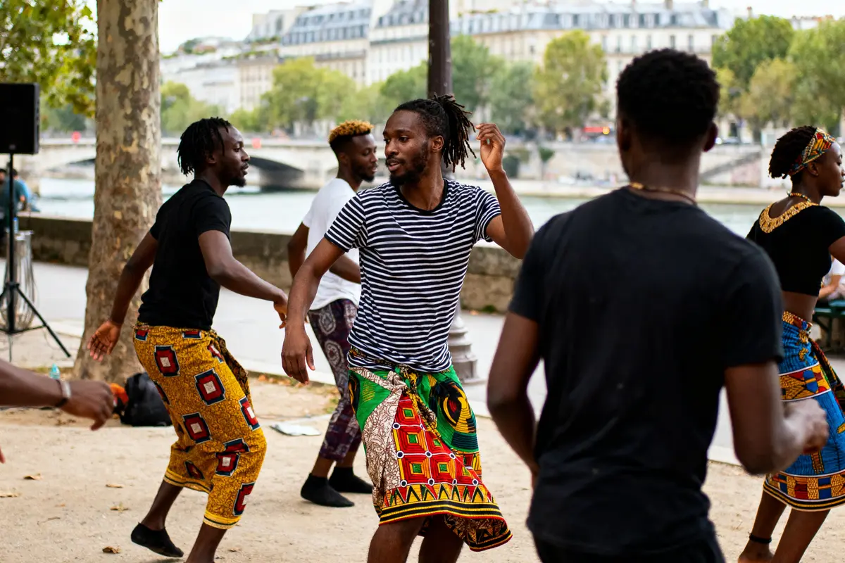 Groupe de personnes dansant en plein air près d'une rivière, portant des vêtements colorés, ambiance joyeuse et festive.