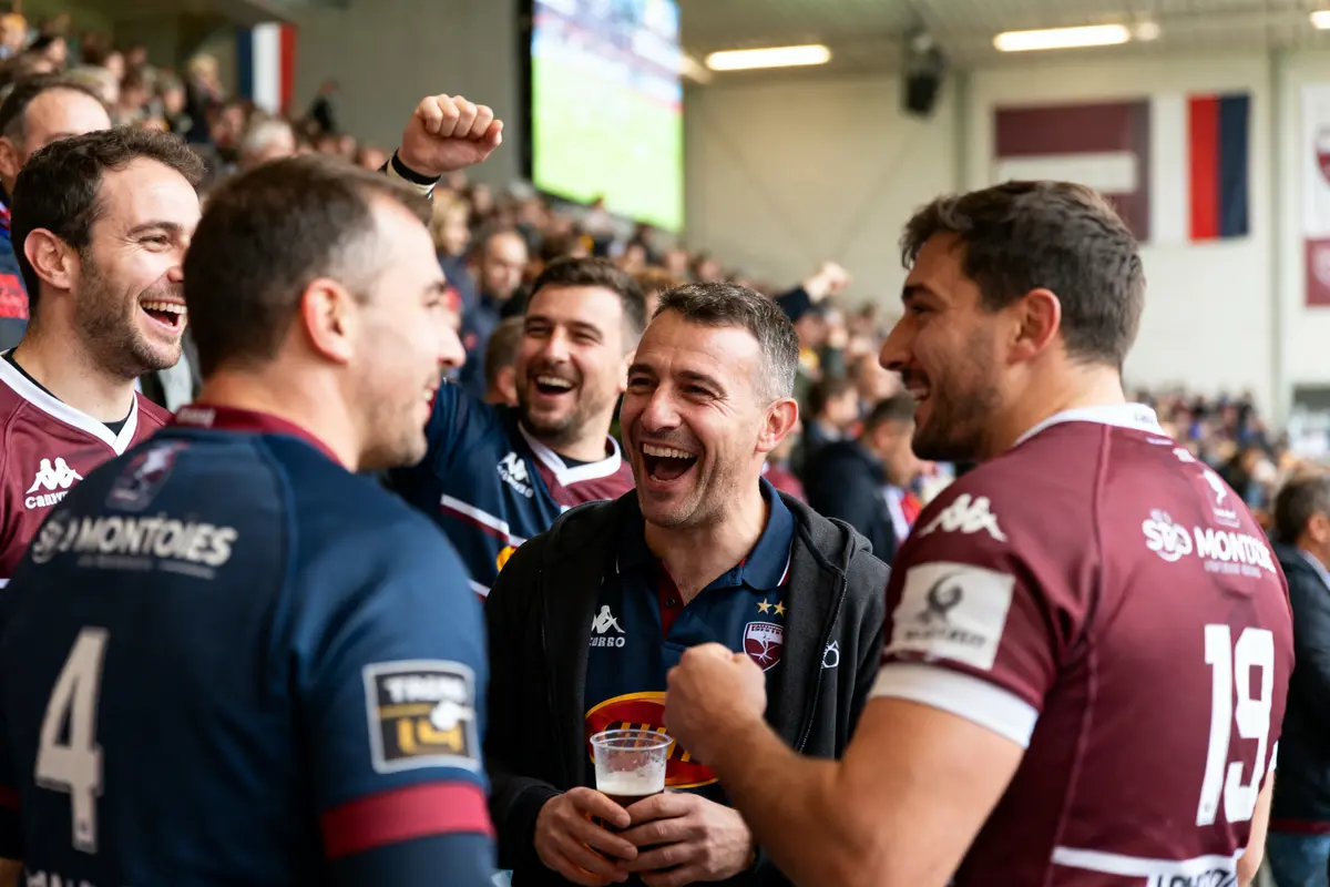 Supporters de rugby souriant dans une tribune de stade, ambiance festive et conviviale autour d'un match.
