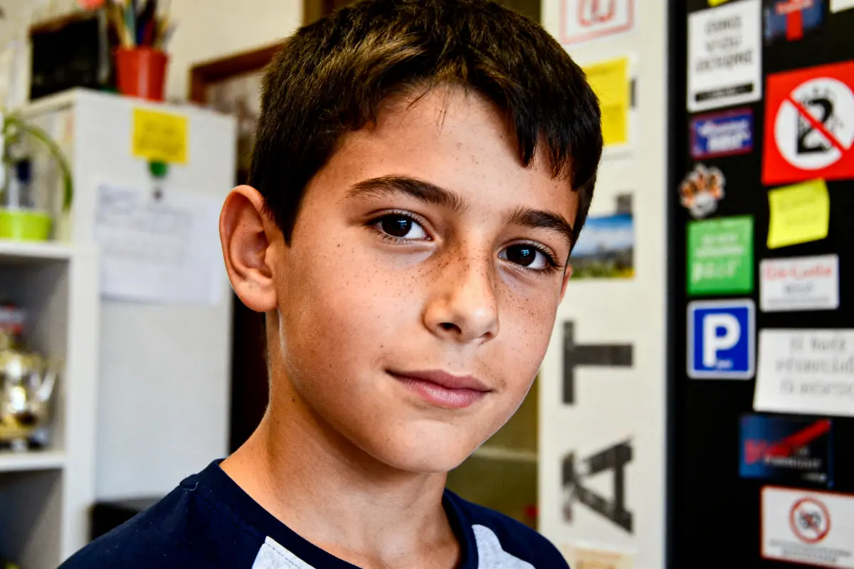 Jeune garçon aux cheveux bruns, regard confiant, dans une pièce avec des affiches et des autocollants colorés.