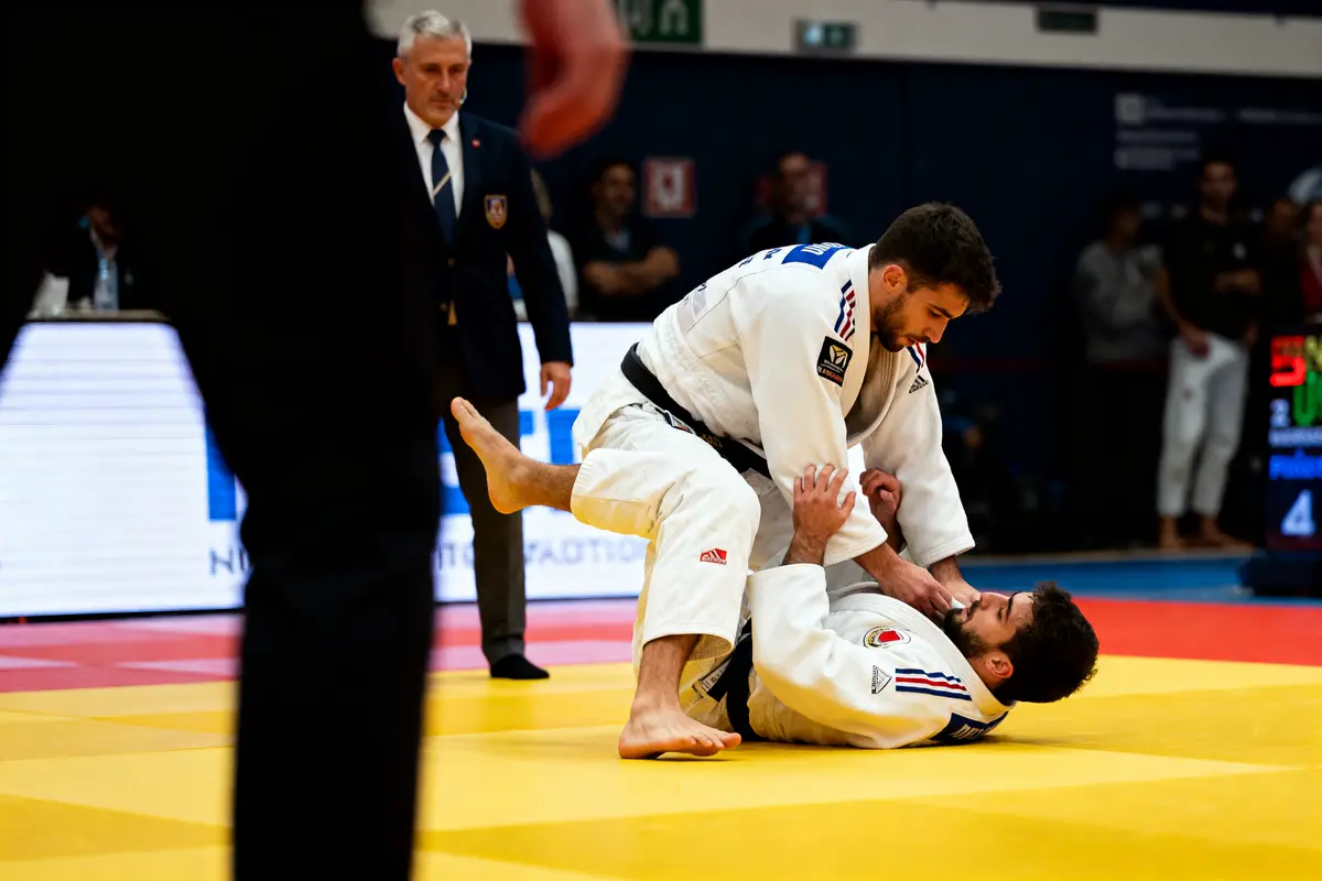 Deux judokas se battent sur un tatami, surveillés par un arbitre en arrière-plan.