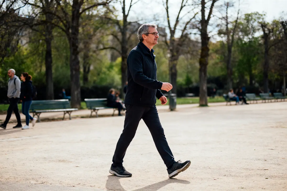 Homme marchant dans un parc, portant des vêtements sombres, arbres et bancs en arrière-plan. Journée ensoleillée.