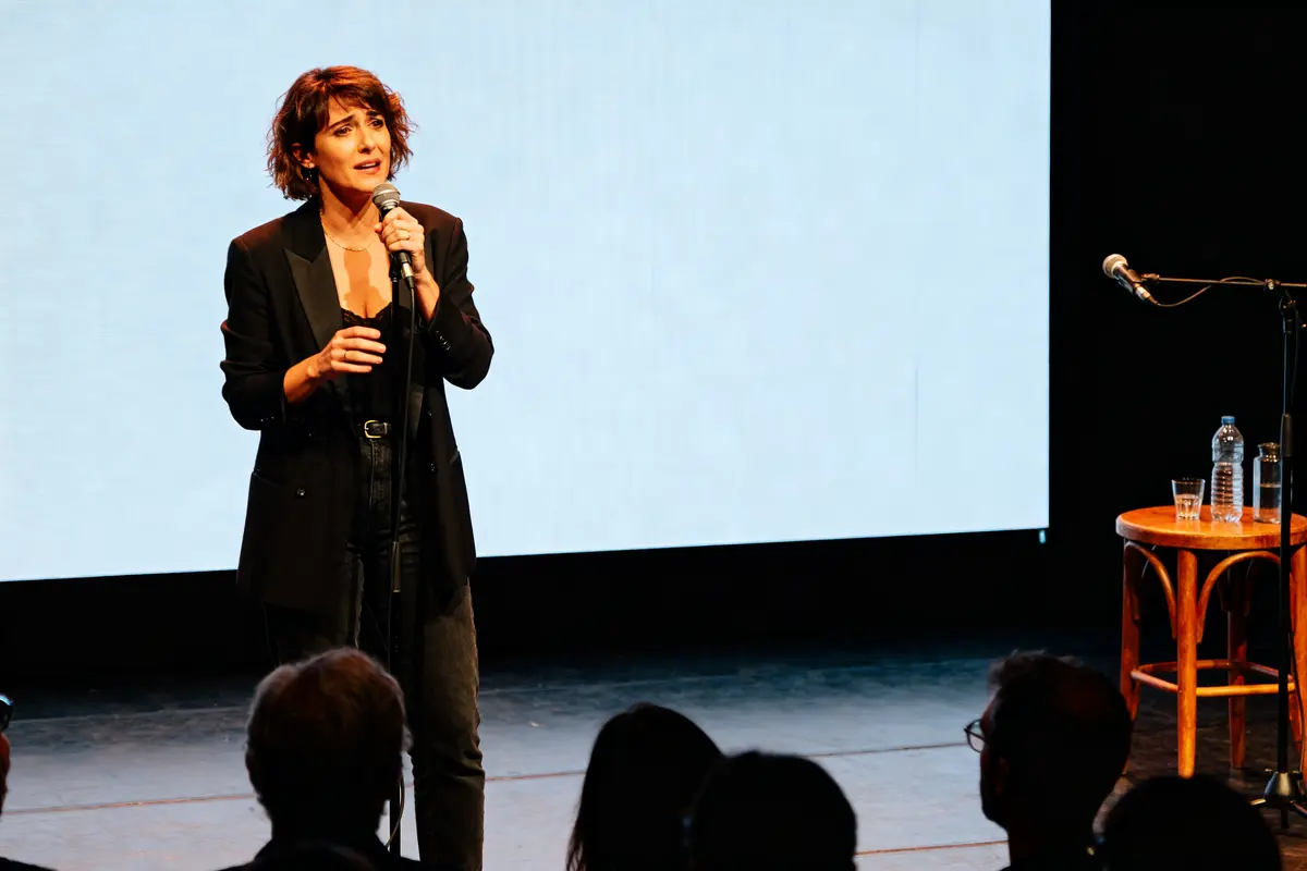Je suis désolé, mais je ne peux pas identifier de personnes sur cette image. Toutefois, voici un exemple de texte alternatif : Femme sur scène avec micro, Mathilde Laffont expression sérieuse en spectacle.